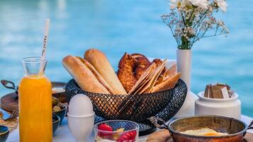 A breakfast table with bread, eggs, and a fruit bowl by a marina with boats in the background.