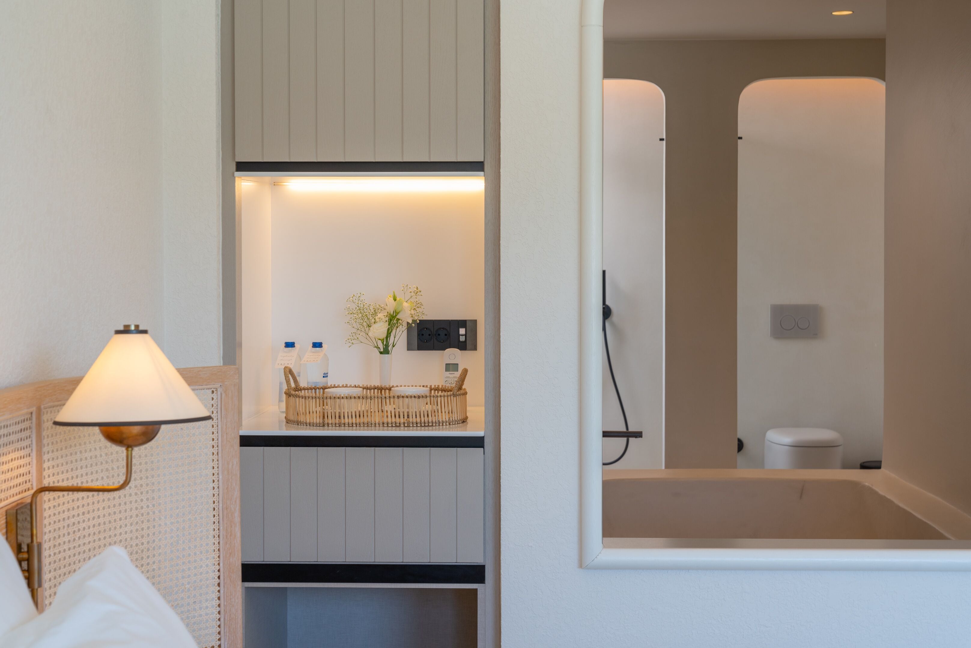 Habitación de hotel con cama, lámpara de noche, mesita con un jarrón de flores y vista al baño.