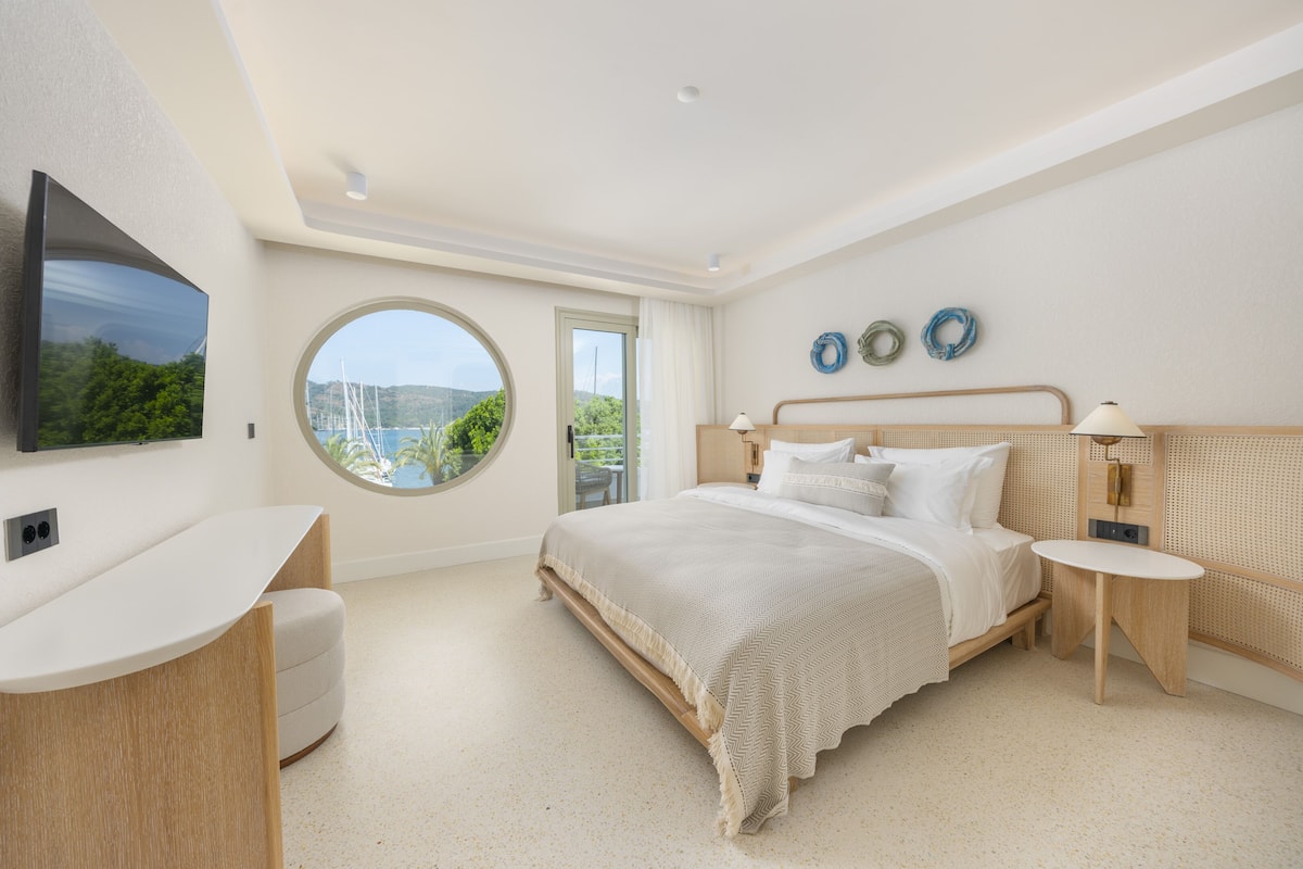 A modern bedroom with a bed, bedside tables, a TV, and a large window offering a view of greenery and boats.