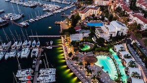 An aerial view of a marina with numerous boats docked, a swimming pool, and surrounding buildings.