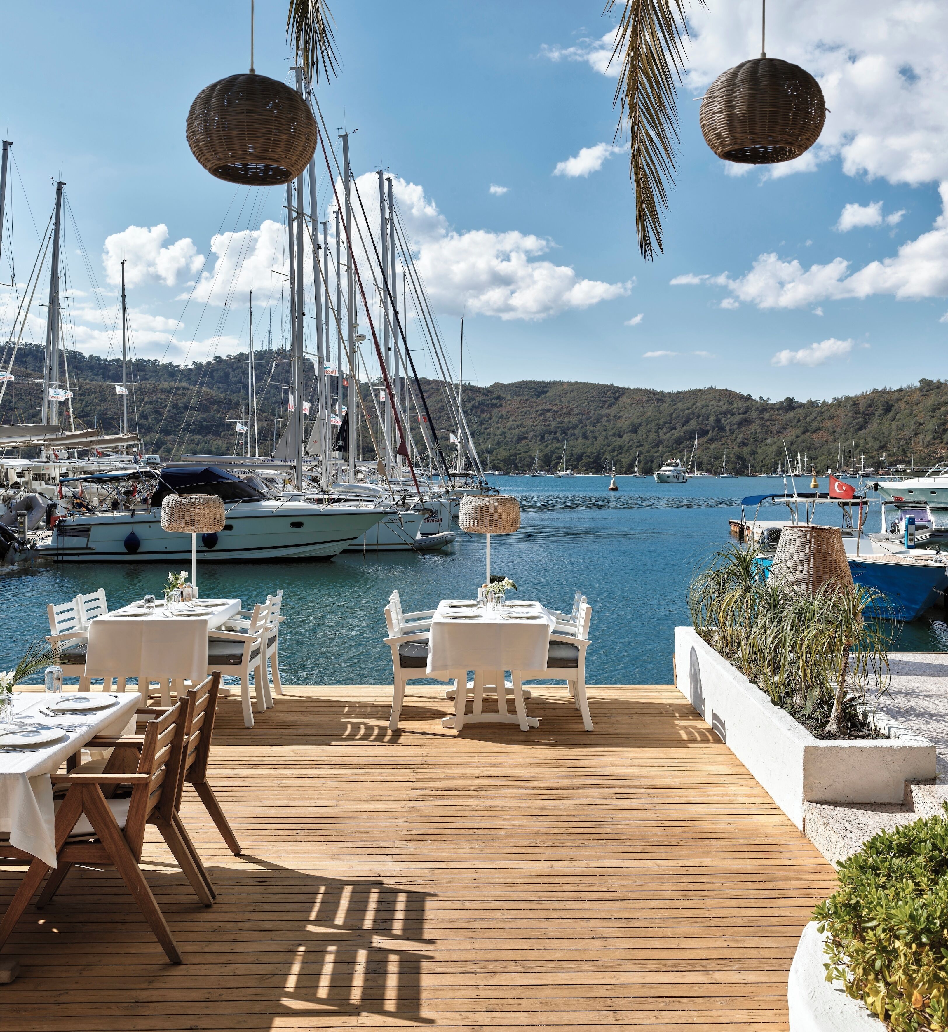 A wooden deck with tables and chairs, overlooking a marina with sailboats and a forested hill in the background.