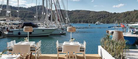 A wooden deck with tables and chairs, overlooking a marina with sailboats and a forested hill in the background.