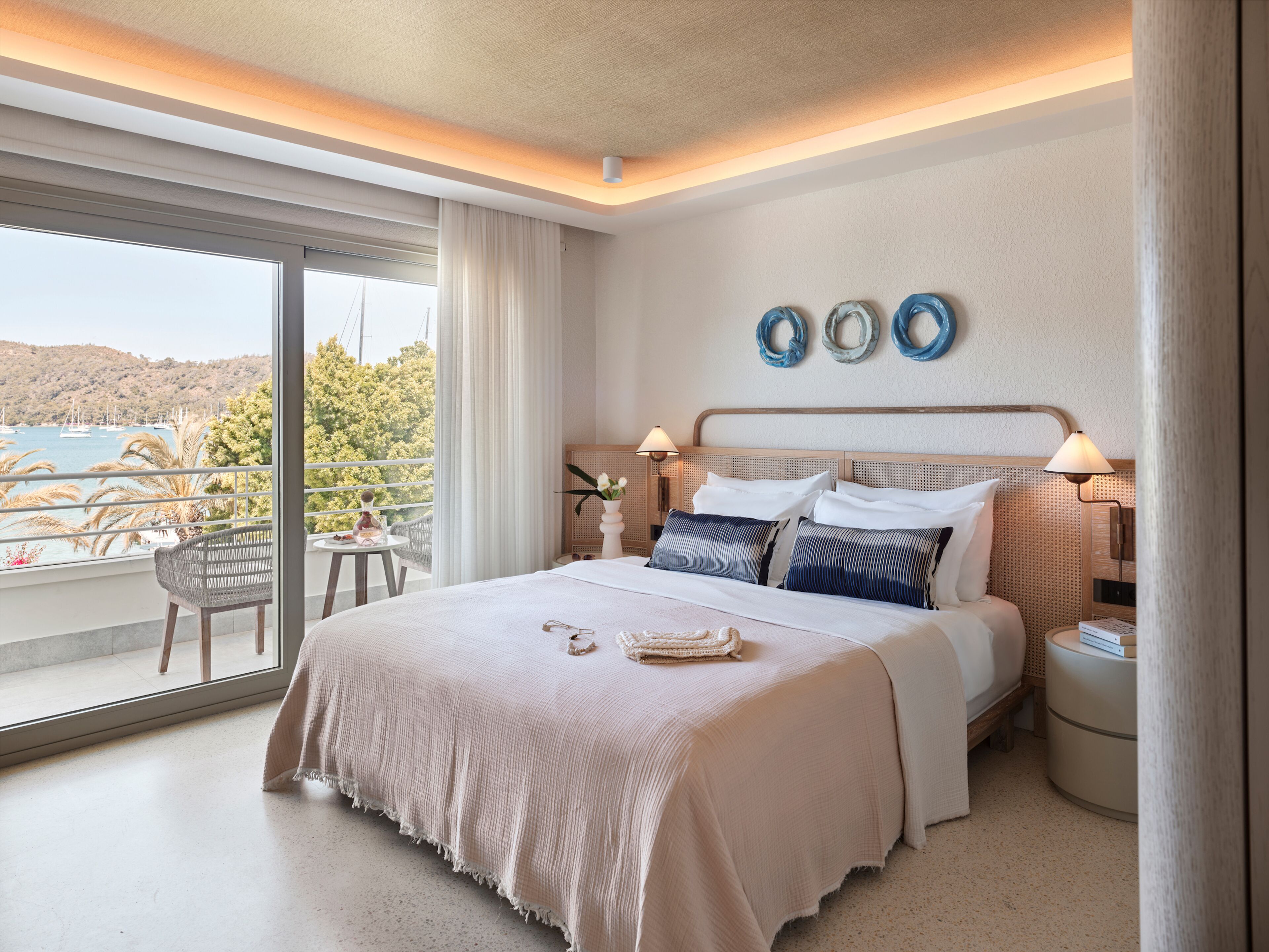 A modern hotel room with a large bed, a balcony with a view of the sea, and a small table with a chair.