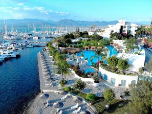 Un port de plaisance avec de nombreux bateaux, une grande piscine entourée de palmiers et un bâtiment portant l’inscription « YACHT ».