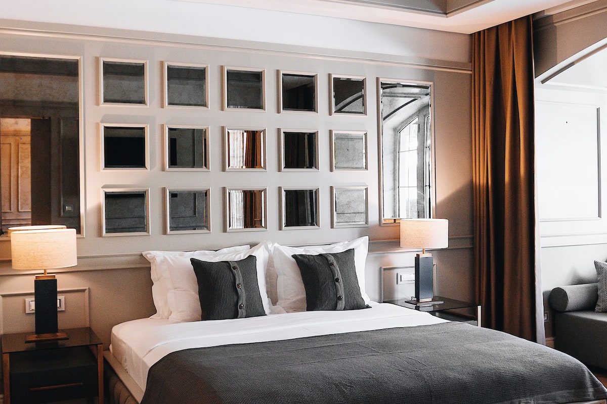 A bedroom with a large bed, a ceiling fan, a bedside table with a lamp, and a wall-mounted shelf with books.