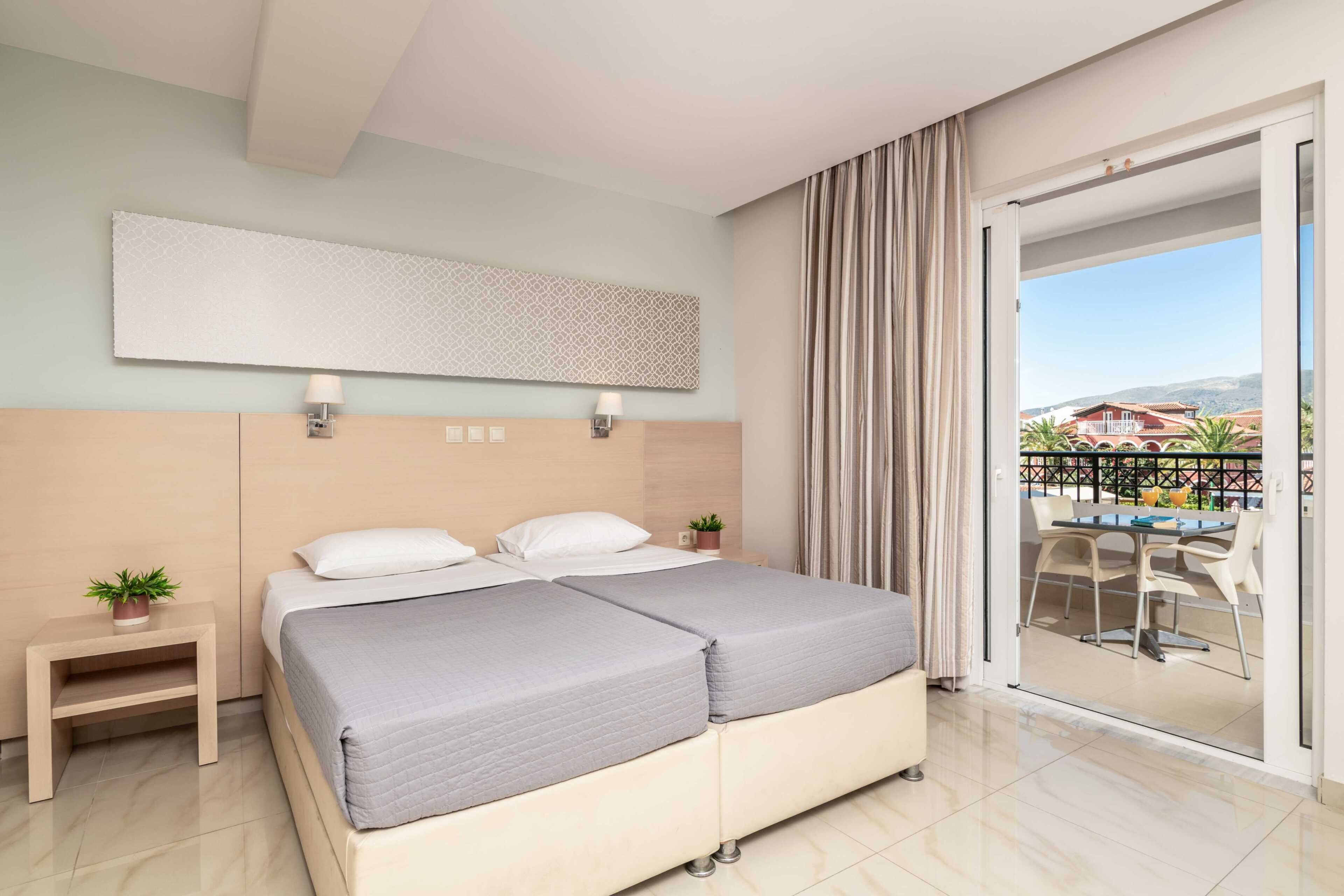 A hotel room with a large bed, a small bedside table with a plant, a wall-mounted light, and a view of the outside balcony.