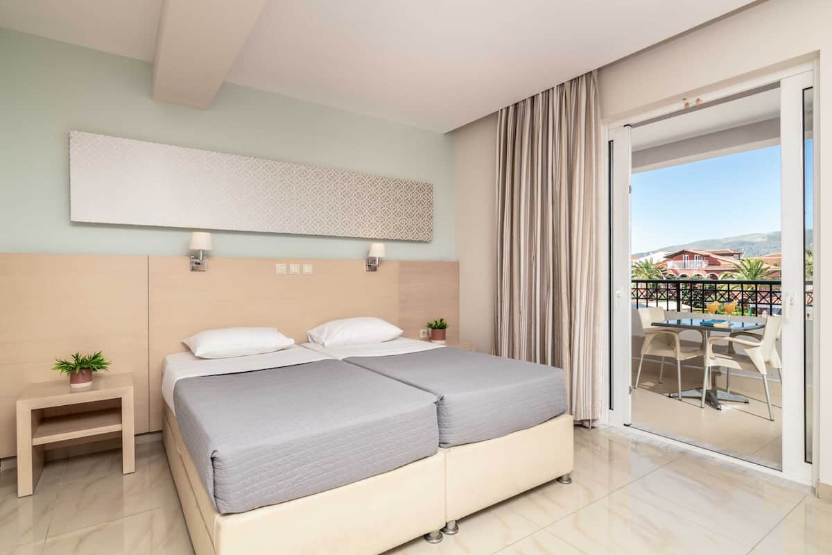 A hotel room with a large bed, a small bedside table with a plant, a wall-mounted light, and a view of the outside balcony.
