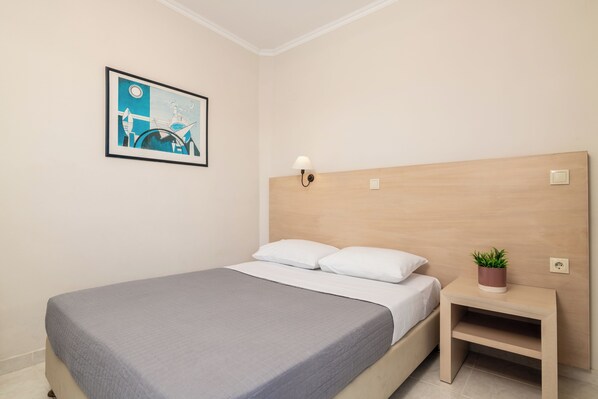 A neatly made bed with a grey bedspread and white pillows, a wooden headboard, a bedside table with a plant, and a framed picture on the wall.