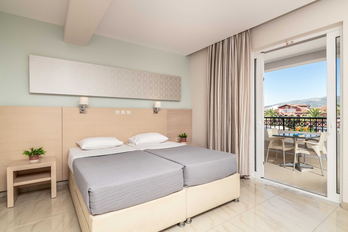 A hotel room with a bed, bedside tables, a view of the outside, and a balcony with a table and chairs.