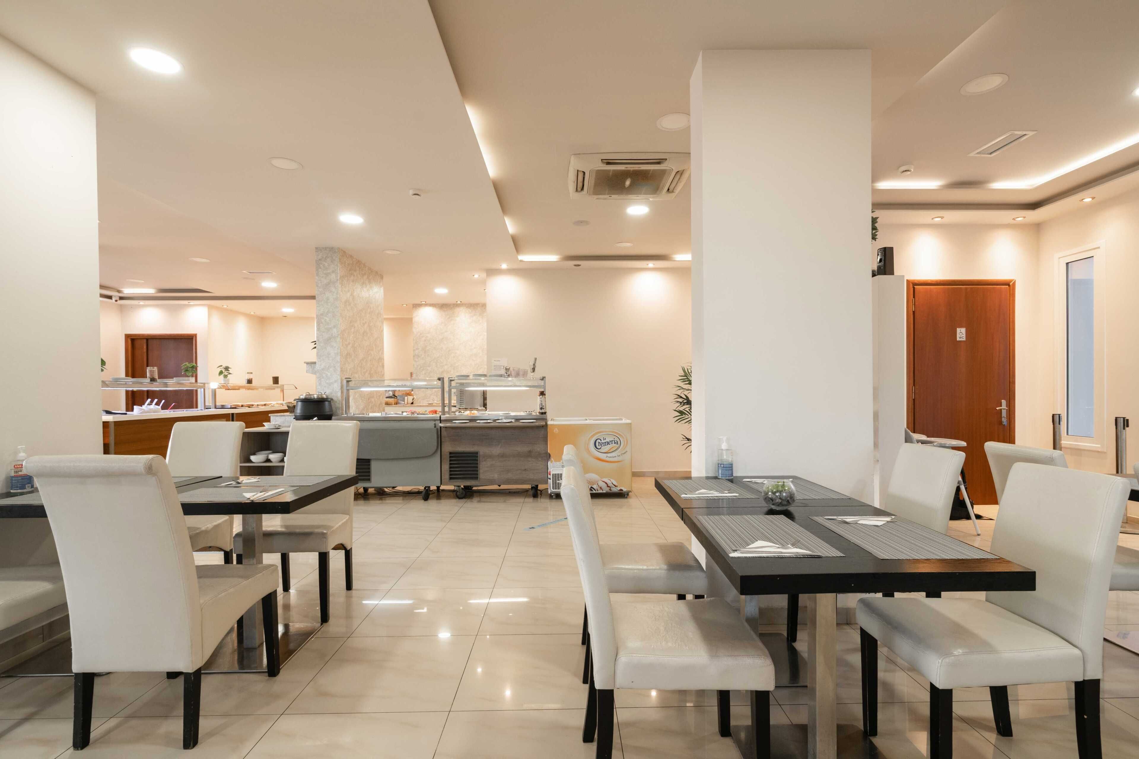 A modern dining area with multiple tables and chairs, a kitchen in the background, and a clean, well-lit environment.