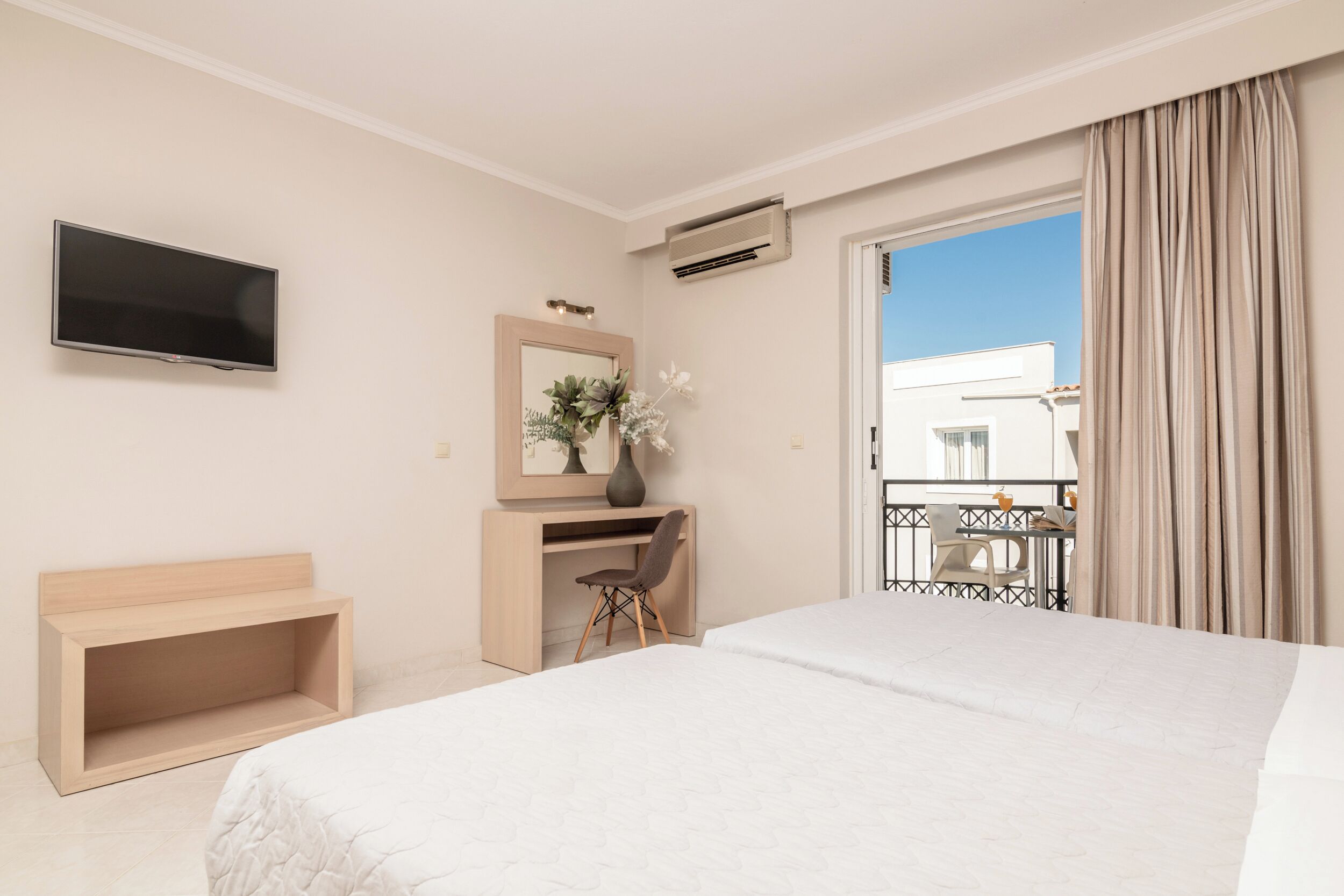 Habitación de hotel con una cama grande, un escritorio con una silla, un televisor de pantalla plana, un espejo y un balcón con vistas a un edificio.