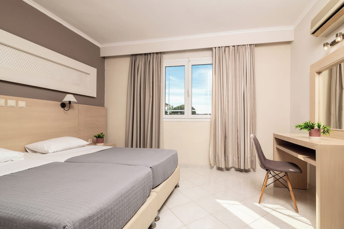 A hotel room with two beds, a desk, a chair, a potted plant, a window with curtains, and an air conditioning unit.