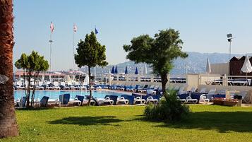 Zona junto a la piscina con sillones de descanso y sombrillas, banderas y vistas a un paisaje urbano de fondo.