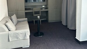 Una habitación de hotel con un sofá gris, una pequeña mesa redonda, un televisor montado en la pared y cortinas grises.