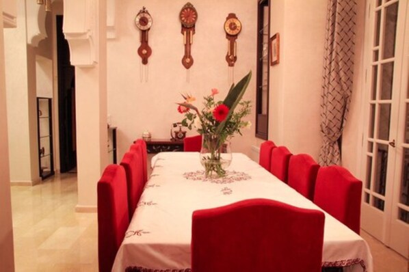 A dining room with a long table, red upholstered chairs, and a vase with flowers on the table. There are three wall clocks above the table. - Dar Chams Tanja (Tangier)