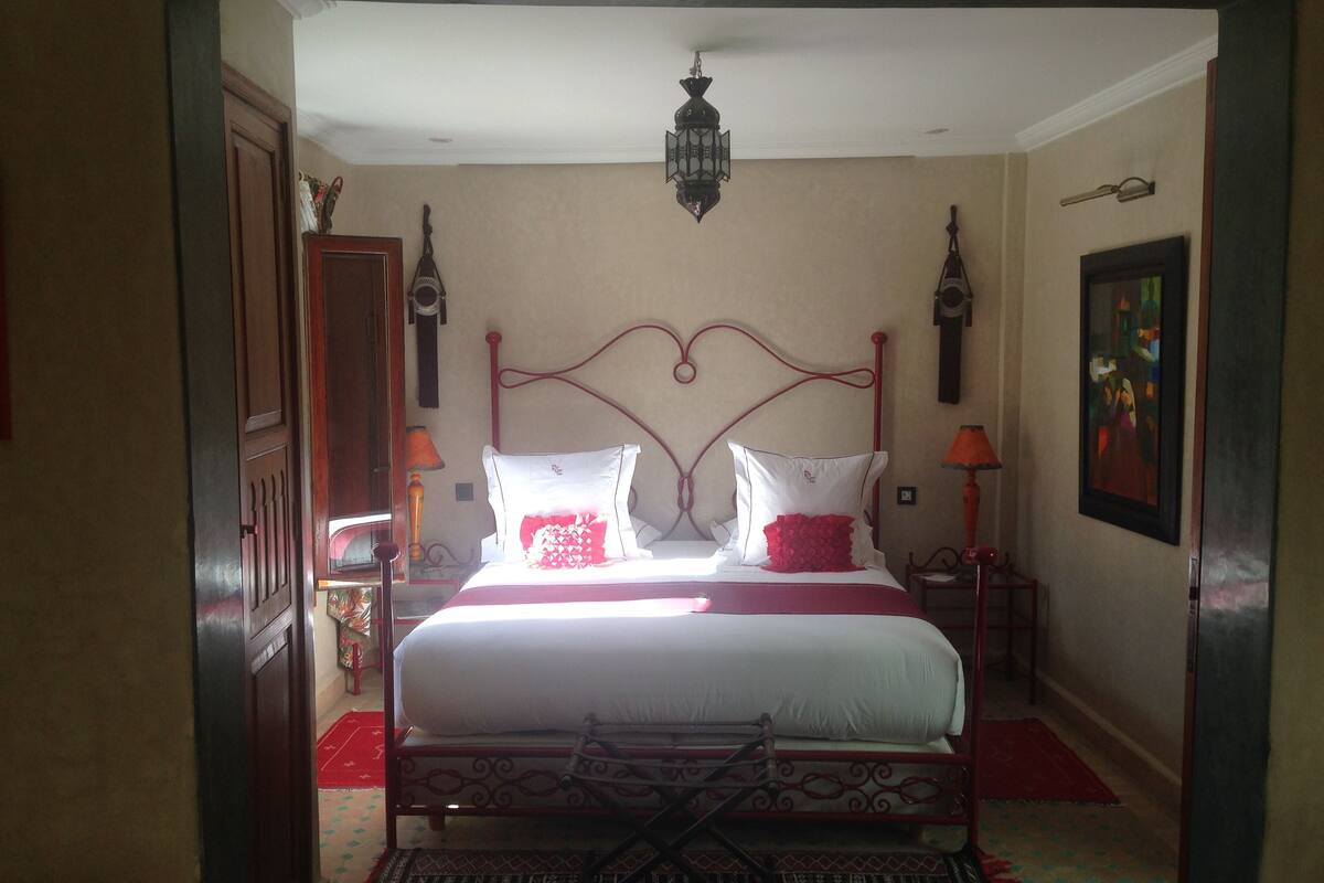 Un dormitorio con cama, dos mesitas de noche, lámpara colgante, un cuadro y una puerta.