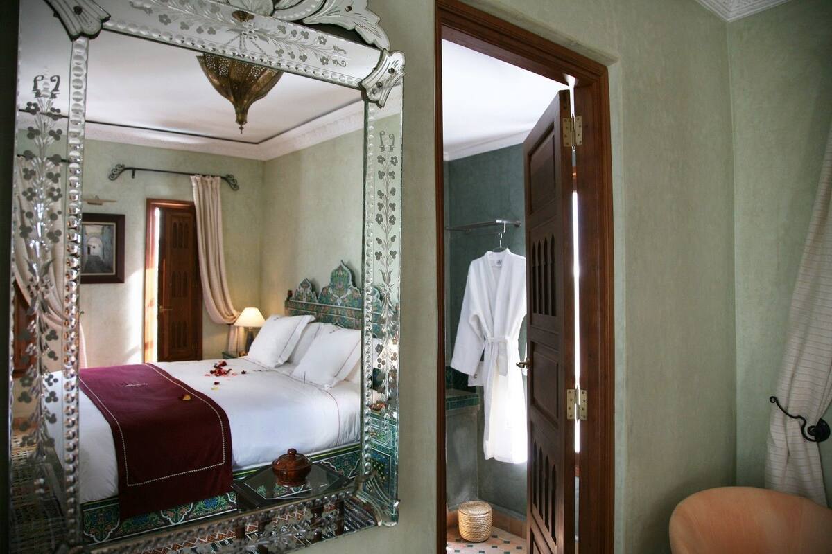 Una habitación con una cama, un espejo grande y decorado, una puerta de madera y una pequeña cesta de mimbre.