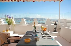 A rooftop terrace with a table set for dining, white outdoor furniture, and a view of the city and hills. - Dar Chams Tanja (Tangier)