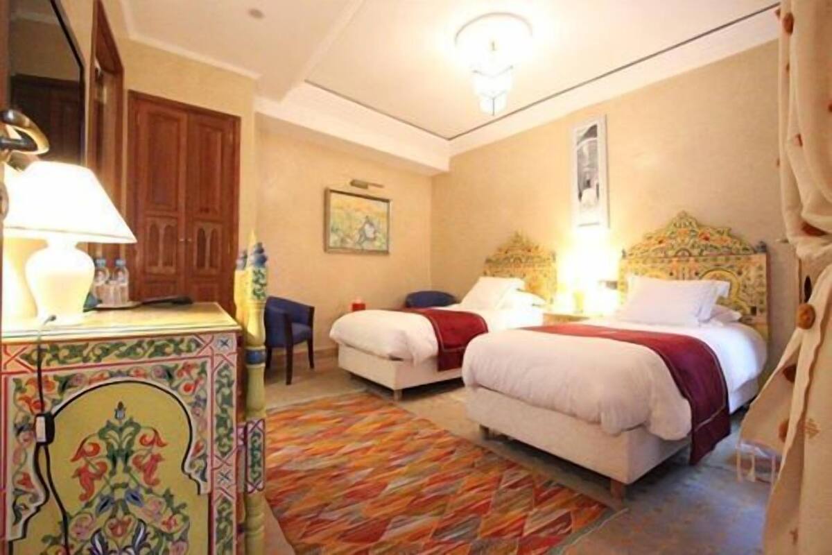 Una habitación de hotel con dos camas, un escritorio con lámpara, una silla y una alfombra colorida.