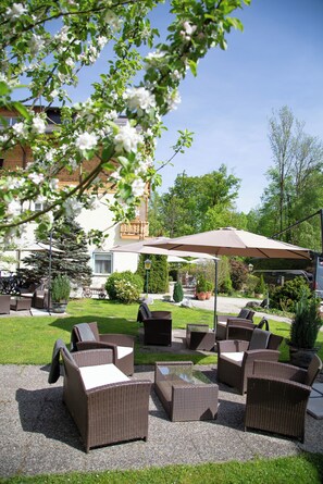 An outdoor seating area with wicker furniture, a covered patio, and a building with a balcony in the background. - Hotel Försterhof (St. Gilgen)