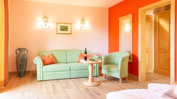 Ein Zimmer mit einem grünen Sofa, einem kleinen runden Tisch mit einer Flasche und Gläsern, einem grünen Sessel, einer großen Vase und Holztüren.