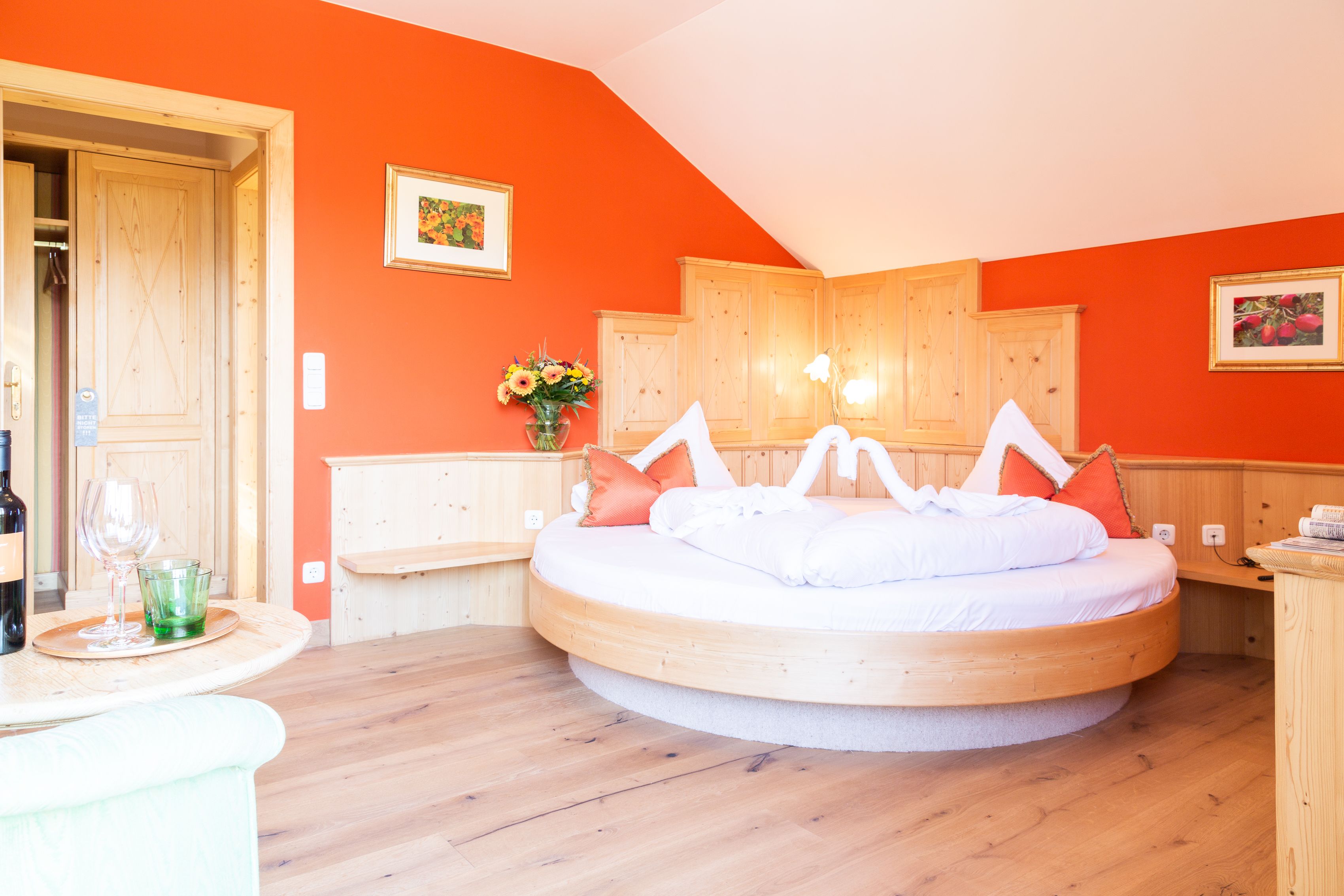 A round bed with white bedding, a wooden headboard, and orange pillows in a room with orange walls and wooden flooring.