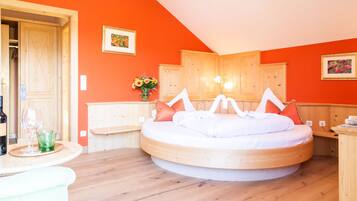 A round bed with white bedding, a wooden headboard, and orange pillows in a room with orange walls and wooden flooring.