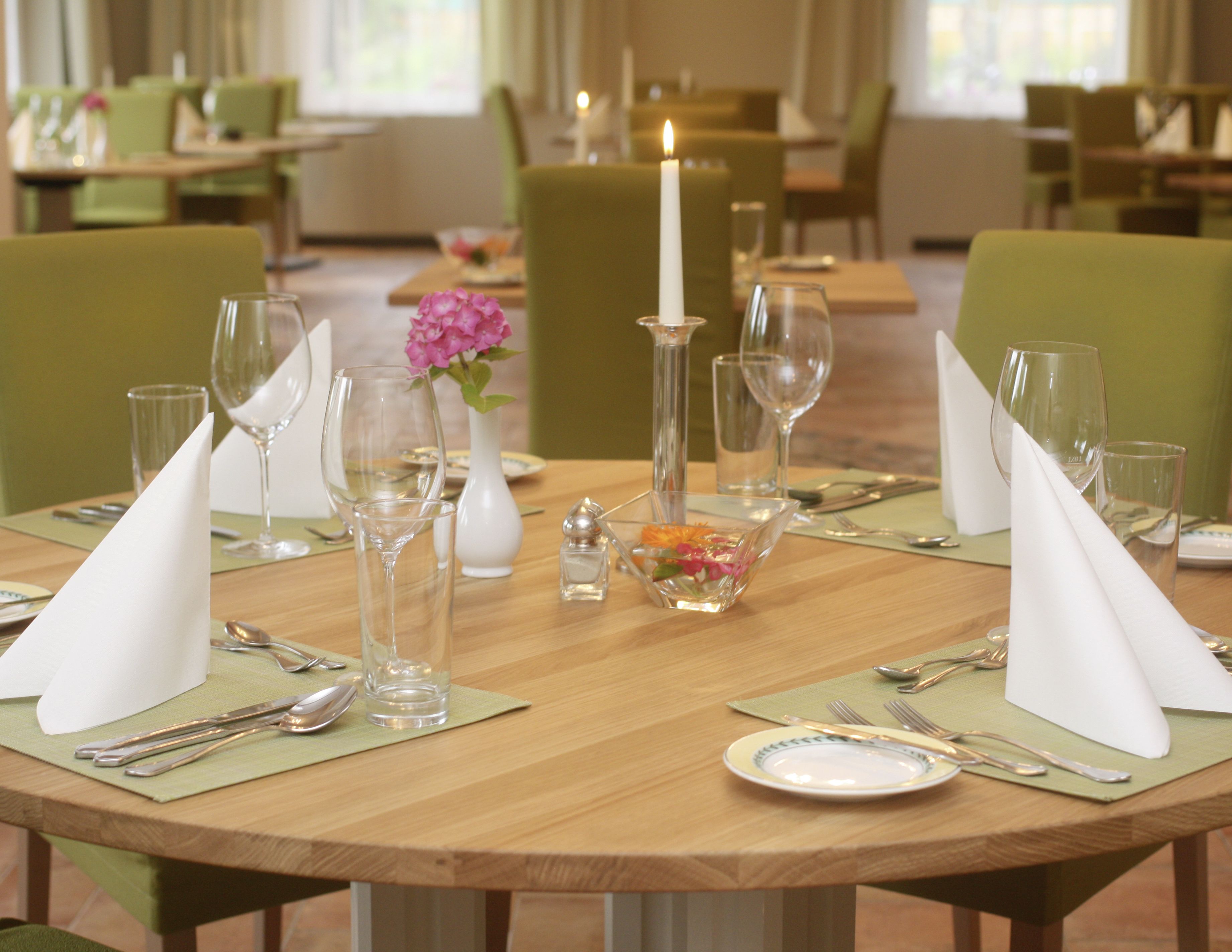 Una mesa de comedor dispuesta con servilletas blancas, copas de vino transparentes, un florero blanco con flores rosadas y una vela encendida.