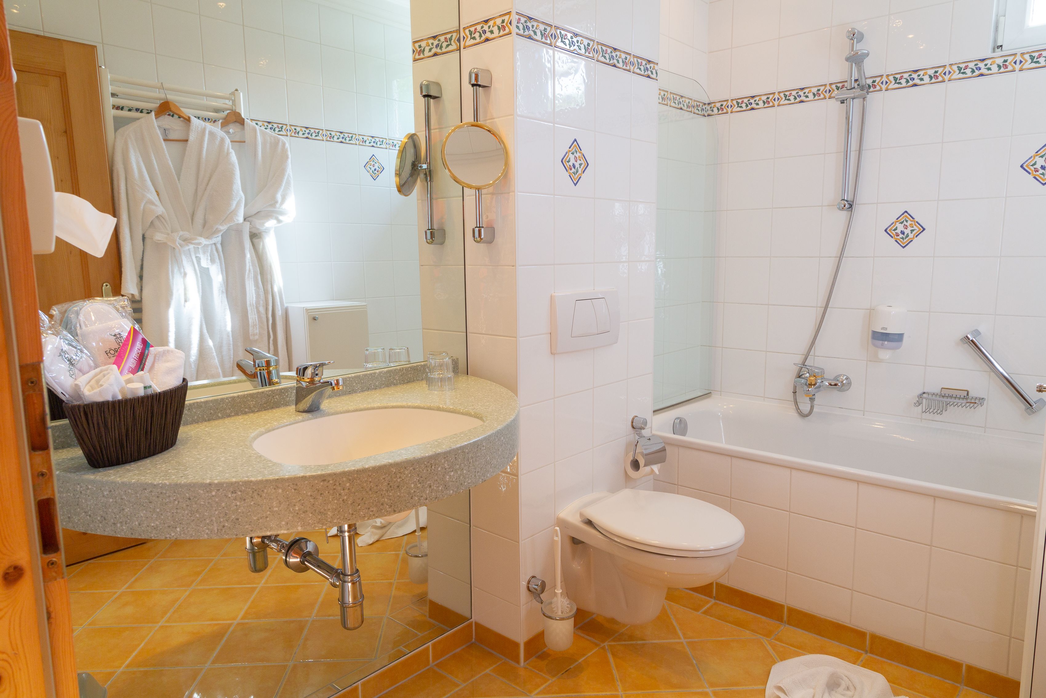 Ein Badezimmer mit Waschbecken, Toilette und Badewanne. Ein Handtuchhalter mit Bademänteln und ein Korb mit Toilettenartikeln sind vorhanden.
