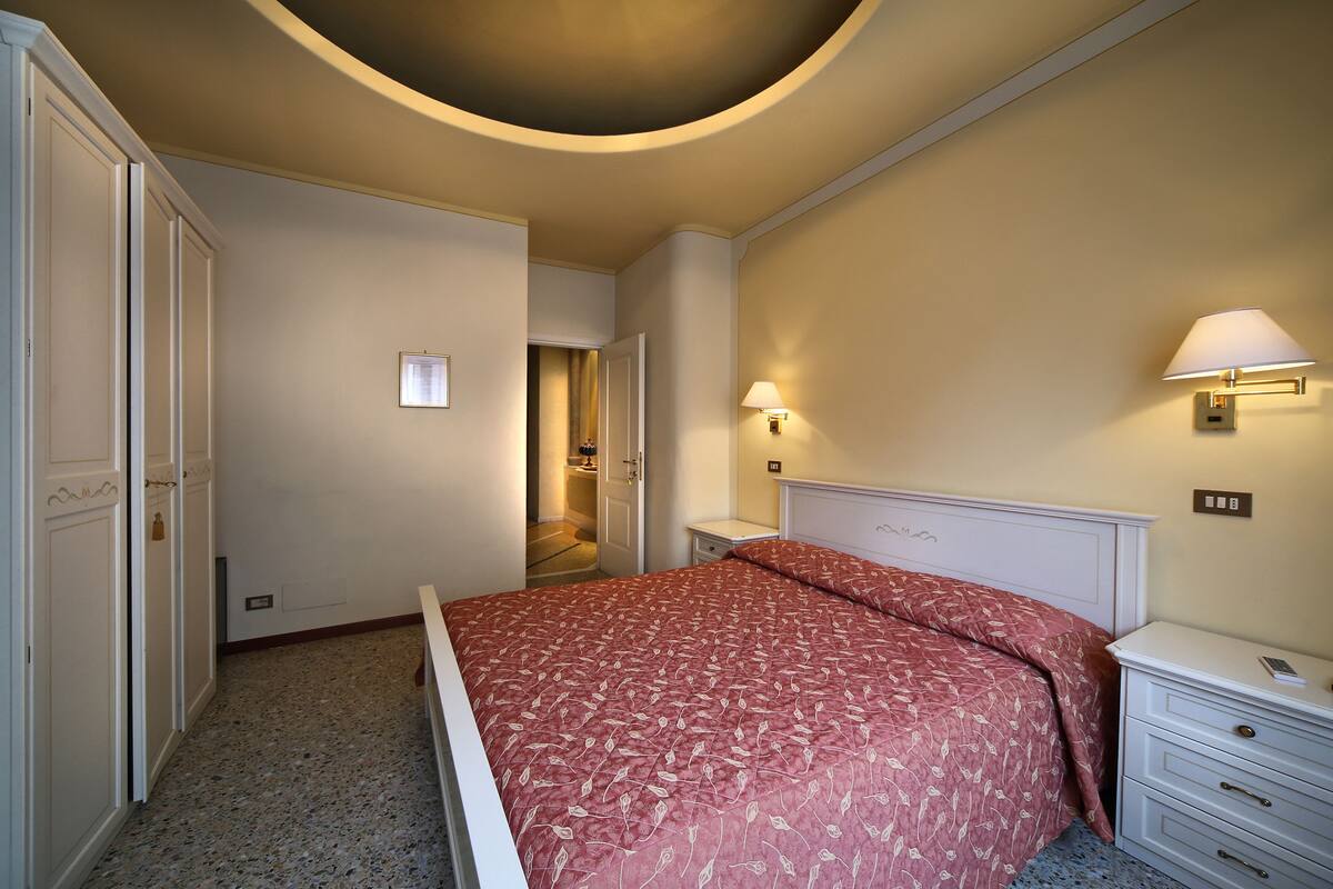 A bedroom with a bed, bedside tables, a nightstand, a lamp, and a door leading to another room.