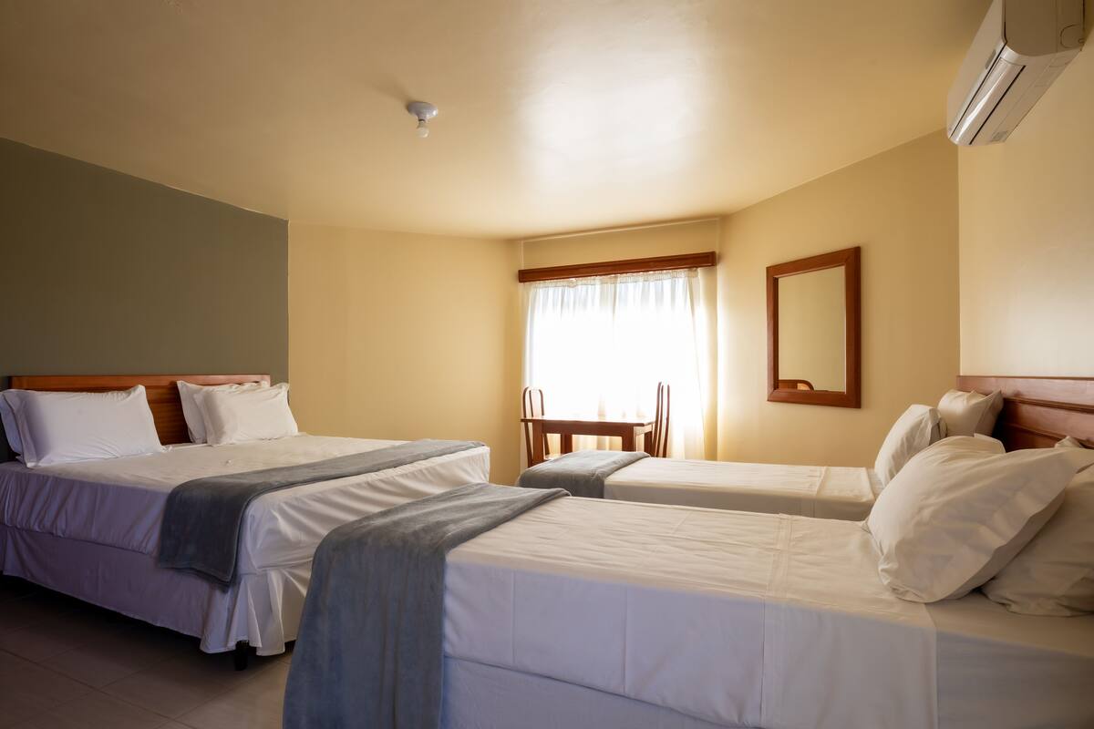A hotel room with two single beds, a wooden headboard, a small table with chairs, a window with curtains, and an air conditioning unit on the wall.