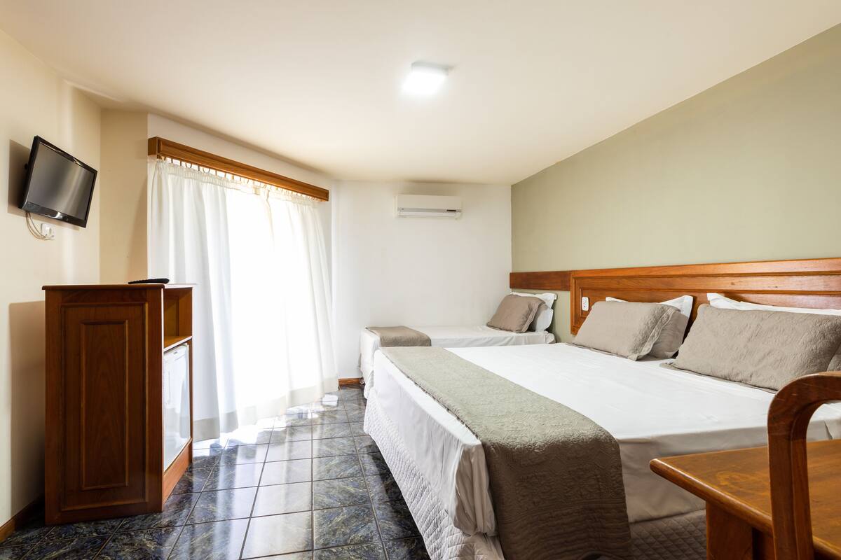 Ein Hotelzimmer mit zwei Betten, einem Holztisch, einem an der Wand befestigten Fernseher und einer Klimaanlage.