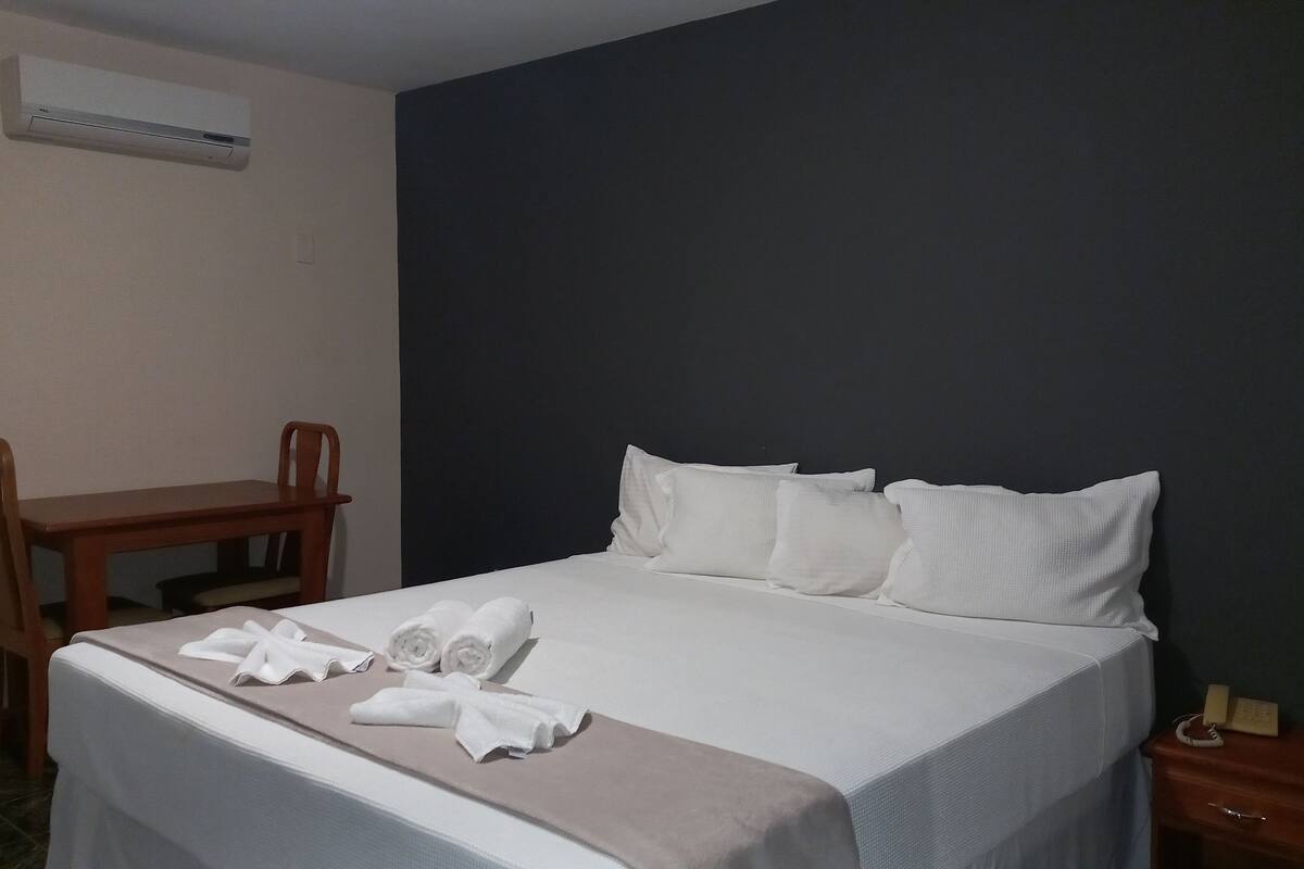 A hotel room with a bed, a wooden chair, a bedside table with a phone, and a wall-mounted air conditioner.