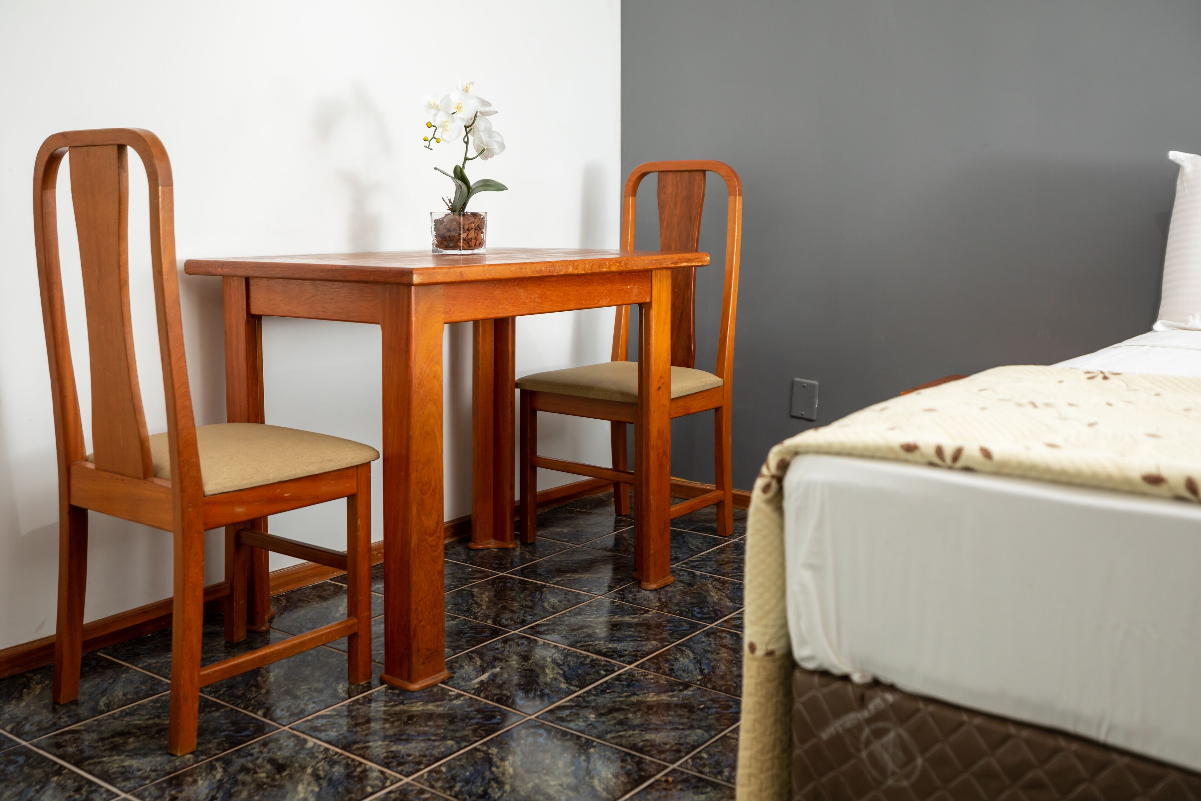 A room with a wooden table and two chairs, a bed with a white bedspread, and a small potted orchid on the table.