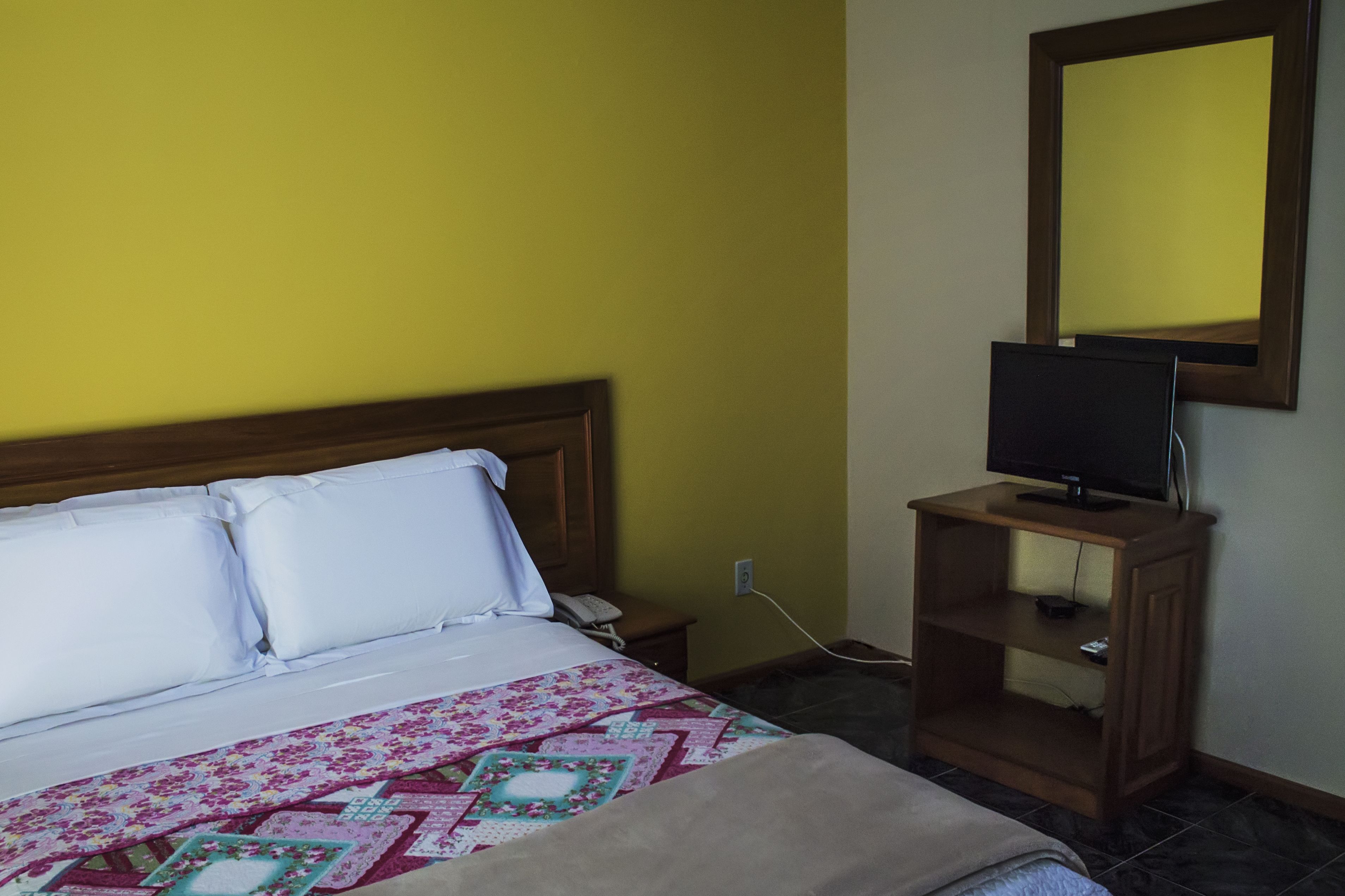 A hotel room with a bed, a bedside table, a television on a wooden stand, and a mirror on the wall.