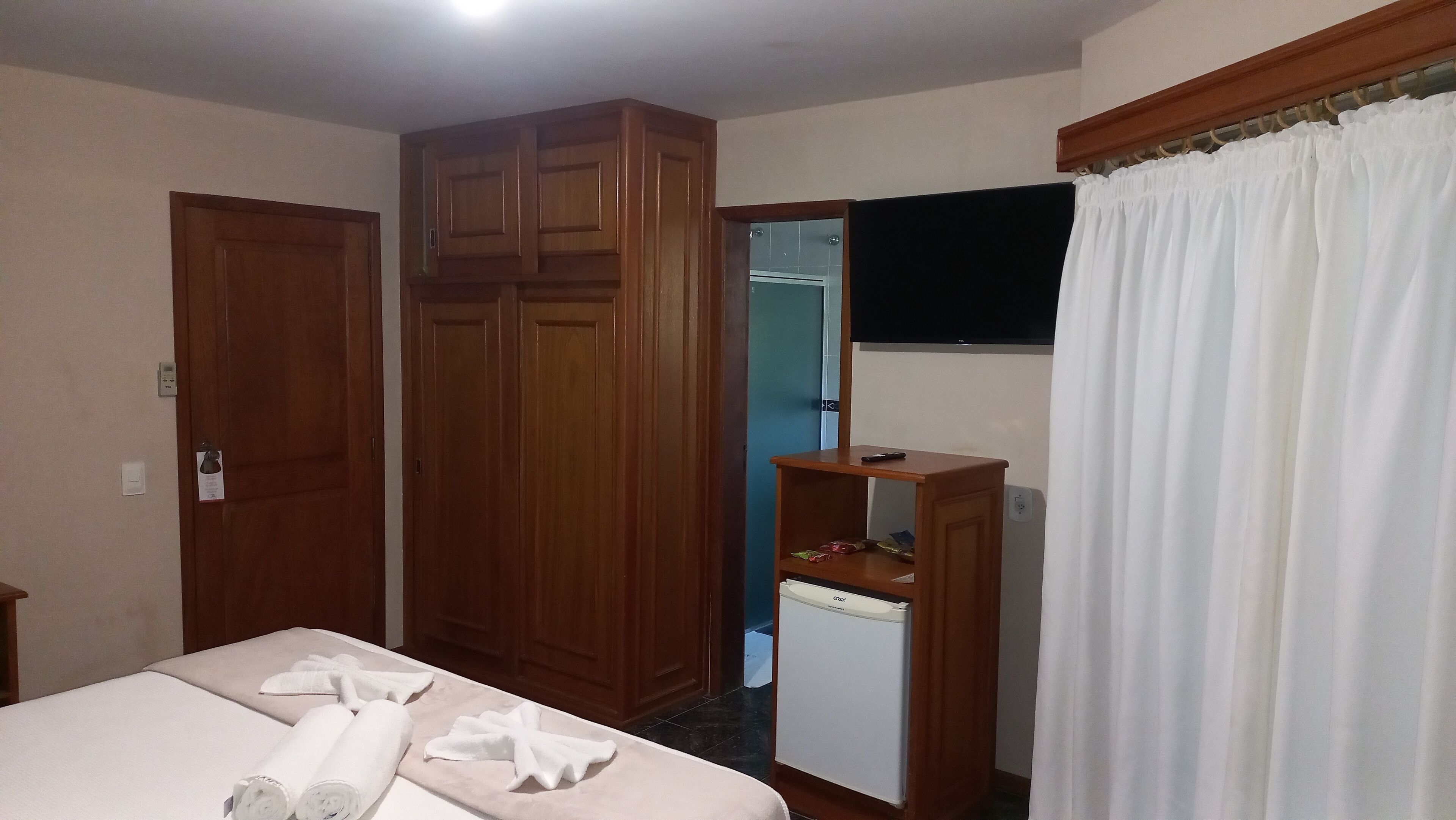 A hotel room with a bed, wooden wardrobe, a small refrigerator, a TV mounted on the wall, and a white curtain.