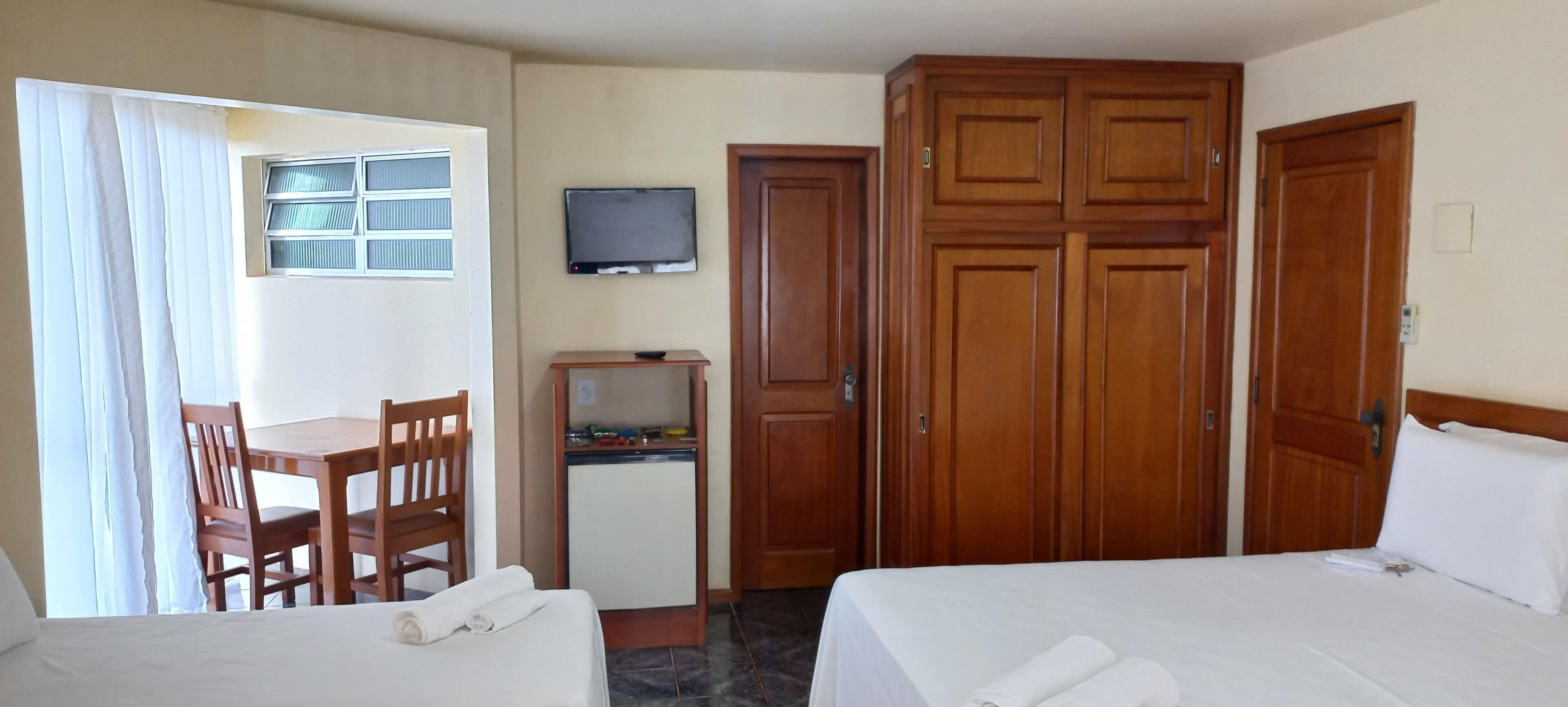 A hotel room with two beds, a desk with a television, wooden wardrobes, and a small kitchenette.