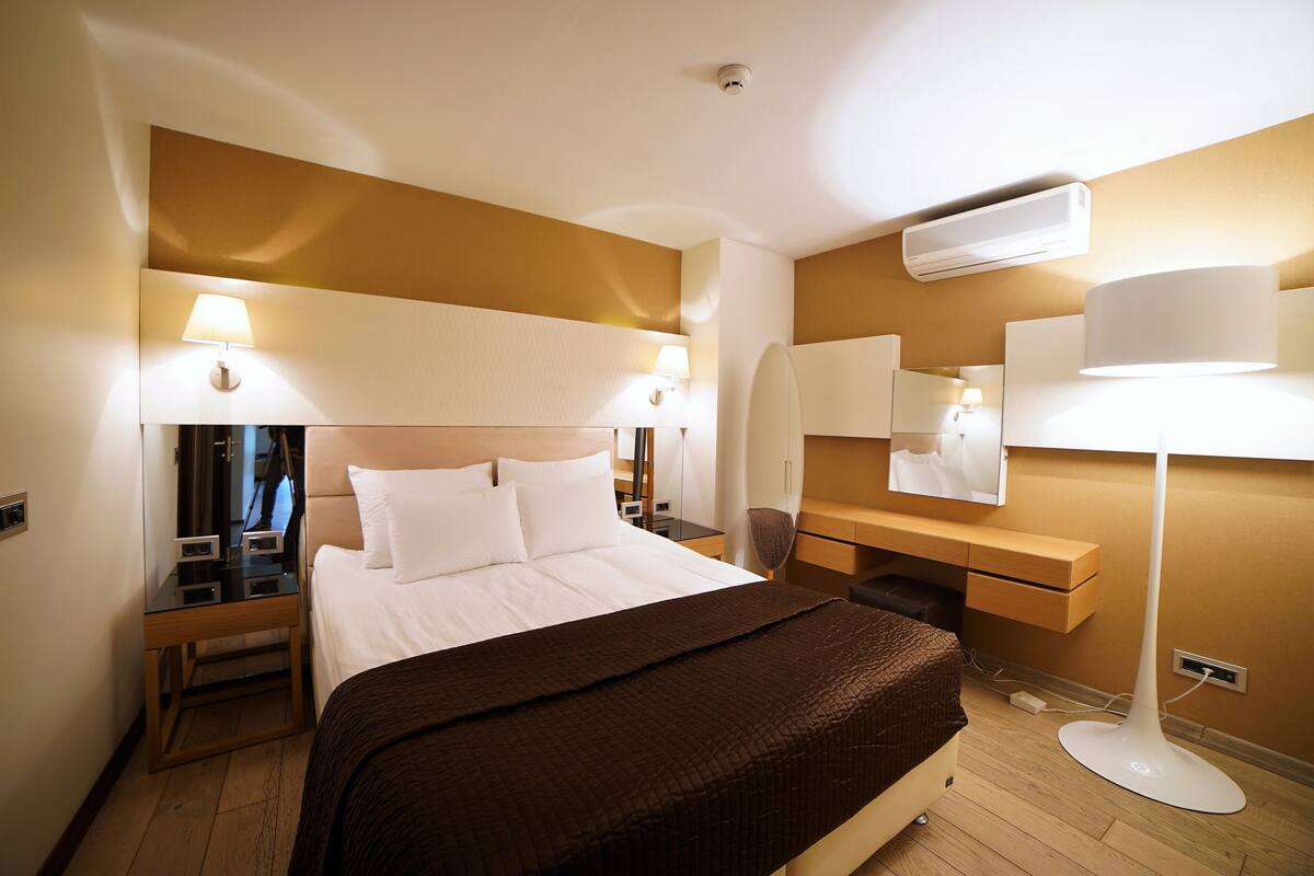 Habitación de hotel con una cama grande, mesitas de noche, un escritorio, una silla, una lámpara, un espejo y un equipo de aire acondicionado.