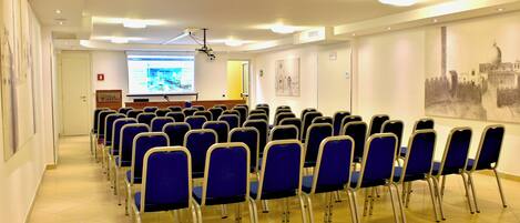 Ein Konferenzraum mit Reihen blauer Stühle, einer Projektionsleinwand und einem Präsentationsbereich.