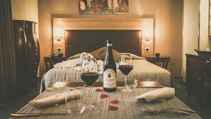 Una cama bien hecha con una botella de vino y copas sobre una mesa, un cuadro en la pared y una lámpara a cada lado de la cama.