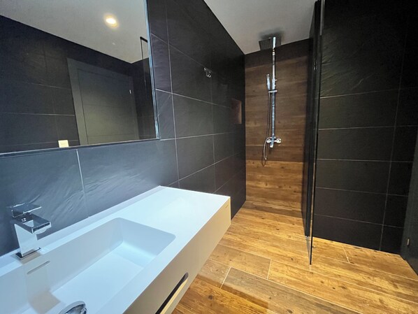 A modern bathroom with a large white bathtub, dark tiled walls, and a wooden floor.