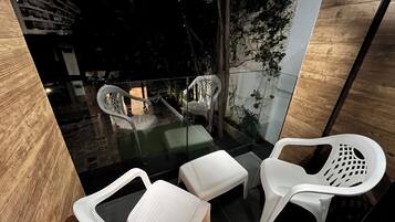 Un área de estar exterior moderna con sillas de plástico blanco y una mesa de vidrio, rodeada por una barandilla de cristal y vistas a un edificio y árboles.