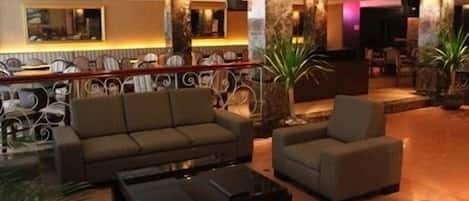 A lobby with a sofa, armchair, and coffee table, with decorative pillars and a reception desk in the background.