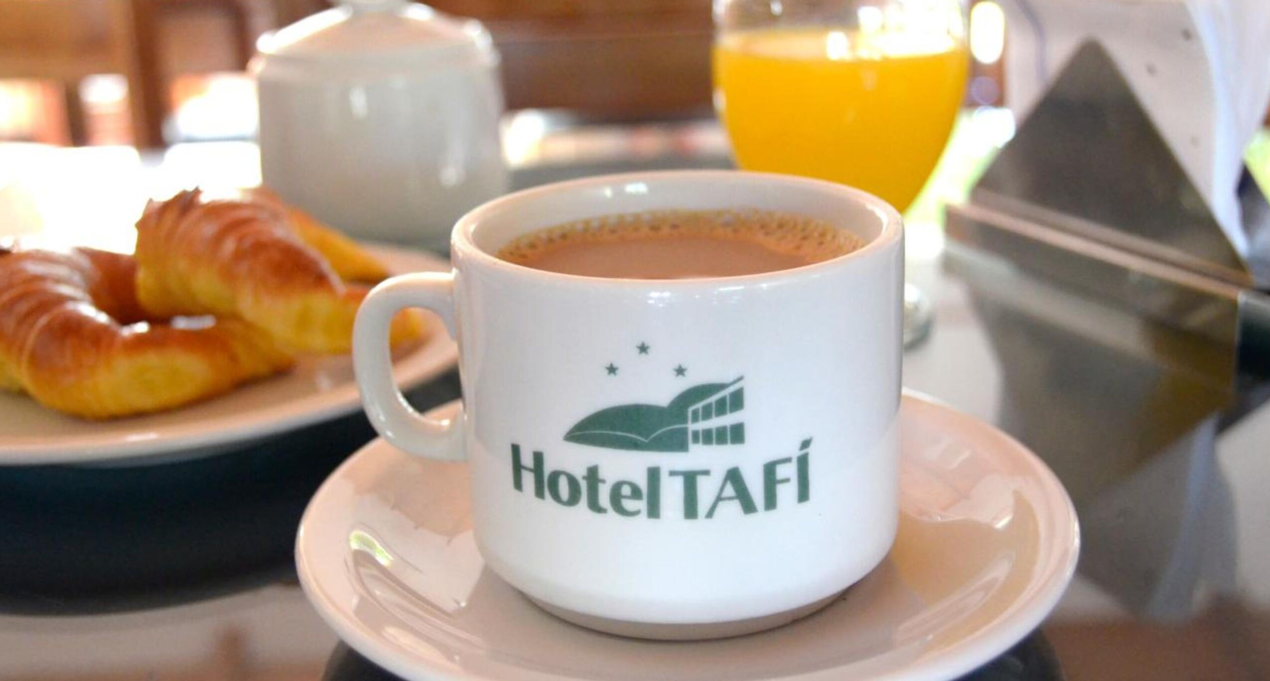 A white HotelTafí mug with a logo, a croissant, a teapot, and a glass of orange juice on a table.