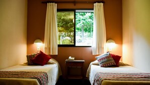 A room with two single beds, each with a pillow and a bedside table. There is a window with curtains, and a lamp on each side of the window. - Hotel Tafi (Tafi del Valle)