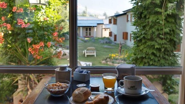 A breakfast table with croissants, a bowl of cereal, a glass of orange juice, and a mug, viewed through a window overlooking a garden and houses. - Hotel Tafi (Tafi del Valle)