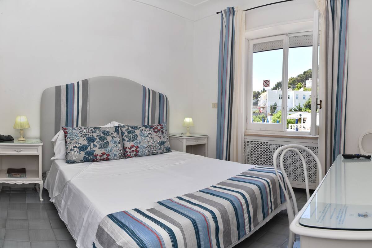 A hotel room with a bed, bedside tables, a TV, a desk, and a window with curtains.