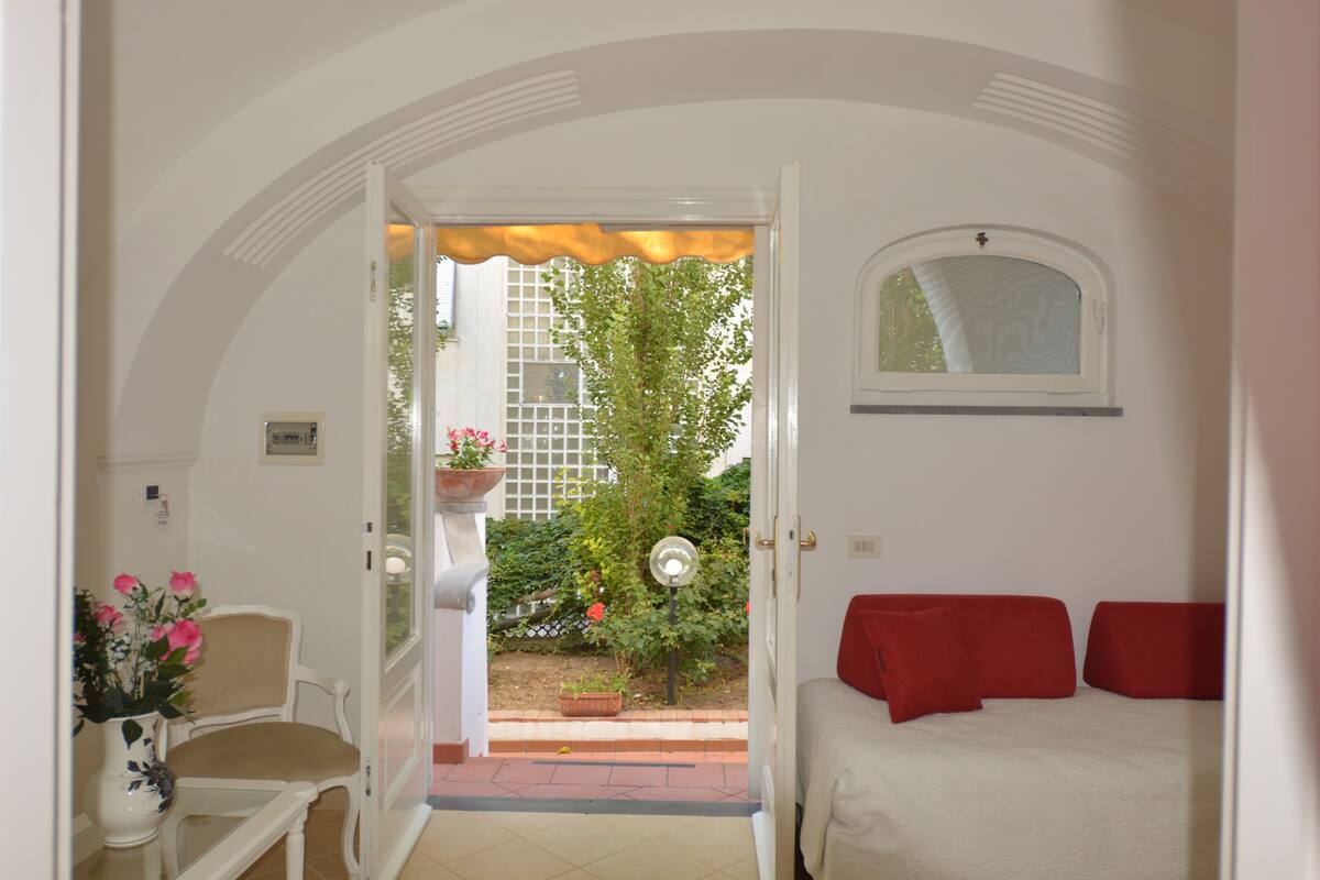 A room with a bed, a chair, a small table with a vase, and a view of a garden through an open door.