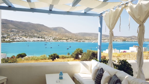 A terrace with a white sofa, a small table, and a view of a coastal town with boats on the water. - Paros Paradise Apartments (Paros)