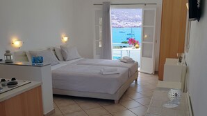 A hotel room with two beds, a balcony with a view of boats and buildings, a small table with a glass, and a TV on the wall. - Paros Paradise Apartments (Paros)